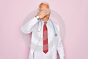 Middle age senior grey-haired doctor man wearing stethoscope and professional medical coat covering eyes with hand, looking