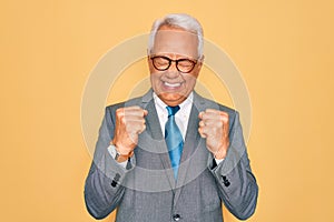 Middle age senior grey-haired handsome business man wearing glasses over yellow background excited for success with arms raised