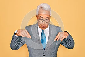 Middle age senior grey-haired handsome business man wearing glasses over yellow background Pointing down with fingers showing
