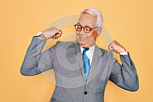 Middle age senior grey-haired handsome business man wearing glasses over yellow background showing arms muscles smiling proud