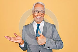 Middle age senior grey-haired handsome business man wearing glasses over yellow background Showing palm hand and doing ok gesture