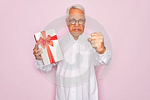 Middle age senior grey-haired man holding romantic gift box over pink background annoyed and frustrated shouting with anger, crazy
