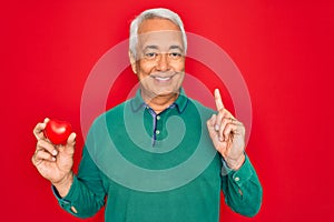 Middle age senior grey-haired man holding romantic red heart object over isolated red background surprised with an idea or