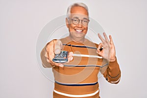 Middle age senior grey-haired man using tv remote control watching television doing ok sign with fingers, excellent symbol