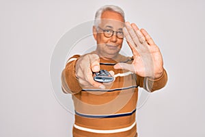 Middle age senior grey-haired man using tv remote control watching television with open hand doing stop sign with serious and