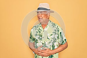 Middle age senior grey-haired man wearing summer hat and floral shirt on beach vacation with hand on stomach because nausea,