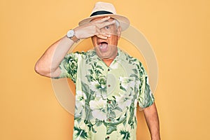 Middle age senior grey-haired man wearing summer hat and floral shirt on beach vacation peeking in shock covering face and eyes