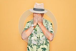 Middle age senior grey-haired man wearing summer hat and floral shirt on beach vacation with sad expression covering face with