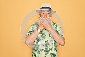 Middle age senior grey-haired man wearing summer hat and floral shirt on beach vacation shocked covering mouth with hands for