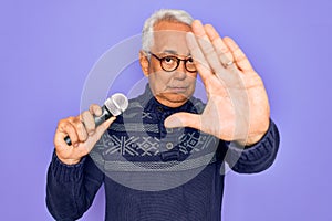 Middle age senior grey-haired singer man singing using music microphone over purple background with open hand doing stop sign with