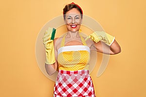 Middle age senior housewife pin up woman wearing 50s style retro dress using cleaner scrub with surprise face pointing finger to