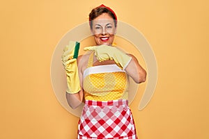 Middle age senior housewife pin up woman wearing 50s style retro dress using cleaner scrub very happy pointing with hand and