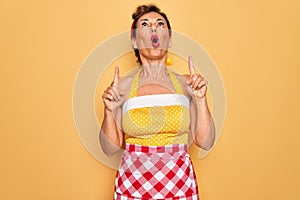 Middle age senior pin up housewife woman wearing 50s style retro dress and apron amazed and surprised looking up and pointing with