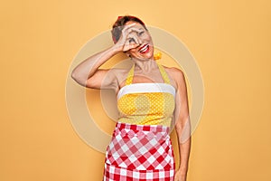 Middle age senior pin up housewife woman wearing 50s style retro dress and apron doing ok gesture with hand smiling, eye looking
