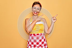 Middle age senior pin up housewife woman wearing 50s style retro dress and apron smiling and looking at the camera pointing with