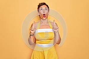 Middle age senior pin up woman wearing 50s style retro dress over yellow background amazed and surprised looking up and pointing