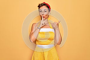 Middle age senior pin up woman wearing 50s style retro dress over yellow background asking to be quiet with finger on lips