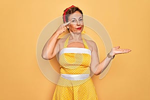 Middle age senior pin up woman wearing 50s style retro dress over yellow background confused and annoyed with open palm showing