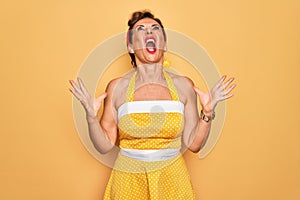 Middle age senior pin up woman wearing 50s style retro dress over yellow background crazy and mad shouting and yelling with