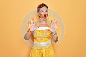 Middle age senior pin up woman wearing 50s style retro dress over yellow background doing stop gesture with hands palms, angry and