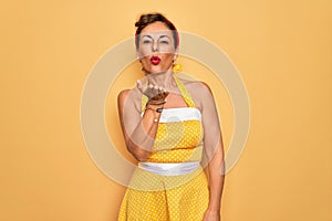 Middle age senior pin up woman wearing 50s style retro dress over yellow background looking at the camera blowing a kiss with hand