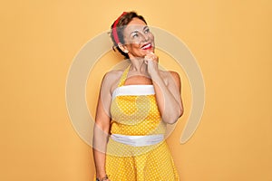 Middle age senior pin up woman wearing 50s style retro dress over yellow background looking confident at the camera smiling with