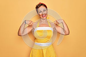 Middle age senior pin up woman wearing 50s style retro dress over yellow background looking confident with smile on face, pointing