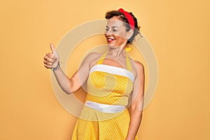 Middle age senior pin up woman wearing 50s style retro dress over yellow background Looking proud, smiling doing thumbs up gesture