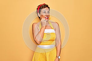 Middle age senior pin up woman wearing 50s style retro dress over yellow background looking stressed and nervous with hands on