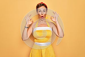 Middle age senior pin up woman wearing 50s style retro dress over yellow background looking surprised and shocked doing ok