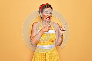 Middle age senior pin up woman wearing 50s style retro dress over yellow background Pointing aside worried and nervous with both