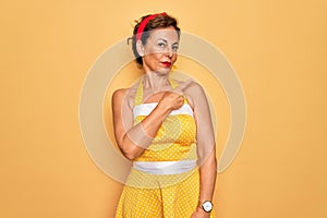 Middle age senior pin up woman wearing 50s style retro dress over yellow background Pointing with hand finger to the side showing