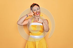 Middle age senior pin up woman wearing 50s style retro dress over yellow background smiling doing talking on the telephone gesture