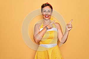 Middle age senior pin up woman wearing 50s style retro dress over yellow background smiling and looking at the camera pointing