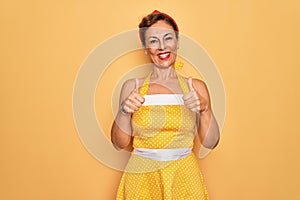 Middle age senior pin up woman wearing 50s style retro dress over yellow background success sign doing positive gesture with hand,