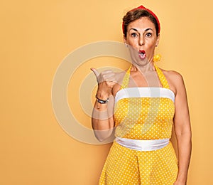 Middle age senior pin up woman wearing 50s style retro dress over yellow background Surprised pointing with hand finger to the