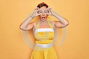 Middle age senior pin up woman wearing 50s style retro dress over yellow background Trying to open eyes with fingers, sleepy and