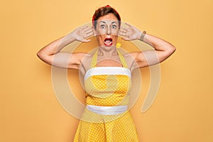 Middle age senior pin up woman wearing 50s style retro dress over yellow background Crazy and scared with hands on head, afraid