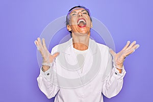 Middle age senior scientist woman wearing coat and laboratory glasses over purple background crazy and mad shouting and yelling