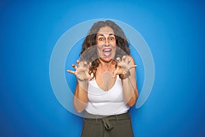 Middle age senior woman with curly hair standing over blue isolated background smiling funny doing claw gesture as cat, aggressive