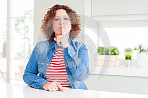 Middle age senior woman with curly hair wearing denim jacket at home asking to be quiet with finger on lips