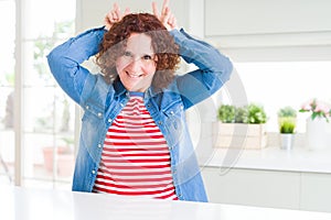 Middle age senior woman with curly hair wearing denim jacket at home Posing funny and crazy with fingers on head as bunny ears,