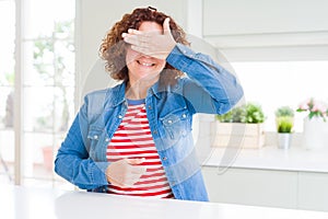 Middle age senior woman with curly hair wearing denim jacket at home smiling and laughing with hand on face covering eyes for