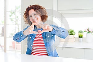 Middle age senior woman with curly hair wearing denim jacket at home smiling in love doing heart symbol shape with hands