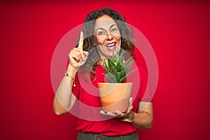 Middle age senior woman holding green cactus over red isolated background surprised with an idea or question pointing finger with