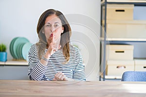 Middle age senior woman sitting at the table at home asking to be quiet with finger on lips