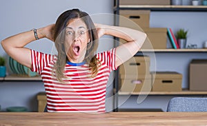 Middle age senior woman sitting at the table at home Crazy and scared with hands on head, afraid and surprised of shock with open