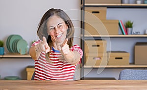 Middle age senior woman sitting at the table at home pointing to you and the camera with fingers, smiling positive and cheerful