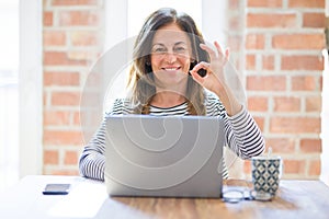 Middle age senior woman sitting at the table at home working using computer laptop doing ok sign with fingers, excellent symbol