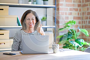 Middle age senior woman sitting at the table at home working using computer laptop happy with big smile doing ok sign, thumb up